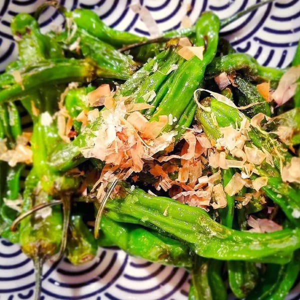 Close-up of green shishito peppers topped with bonito flakes in a patterned bowl.