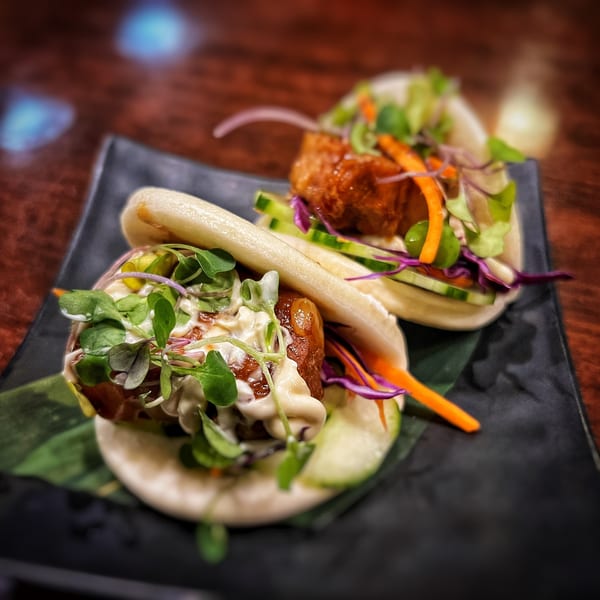 Two bao buns filled with crispy pork belly, fresh greens, and a creamy sauce on a dark plate.
