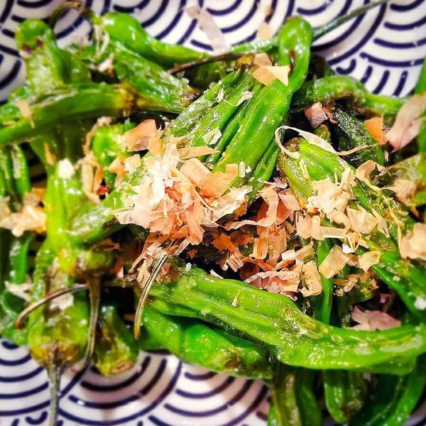Close-up of green shishito peppers topped with bonito flakes in a patterned bowl.
